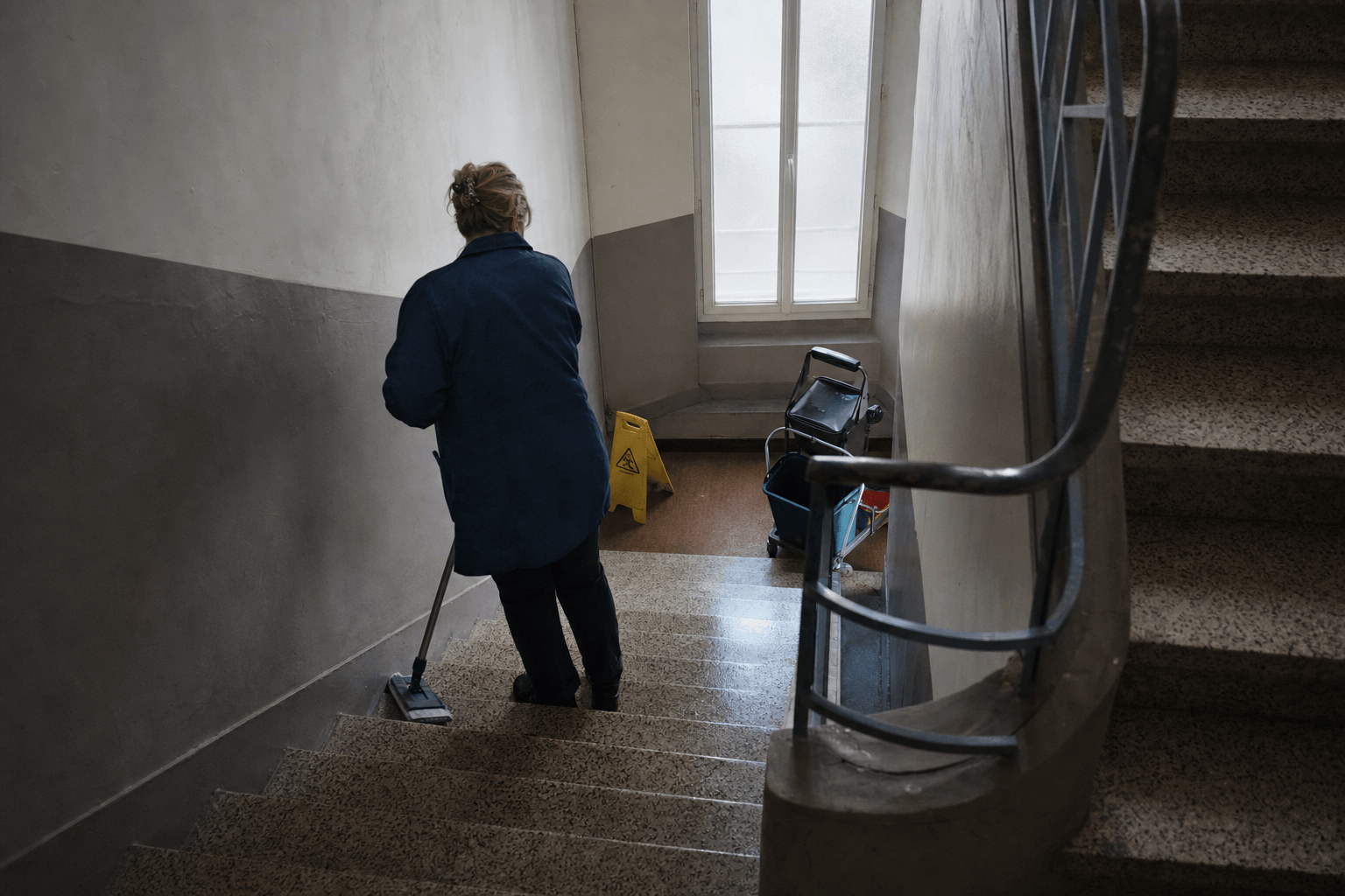 Agent d'entretien de dos lavant les marches d'une cage d'escalier d'immeuble, panneau sol glissant
